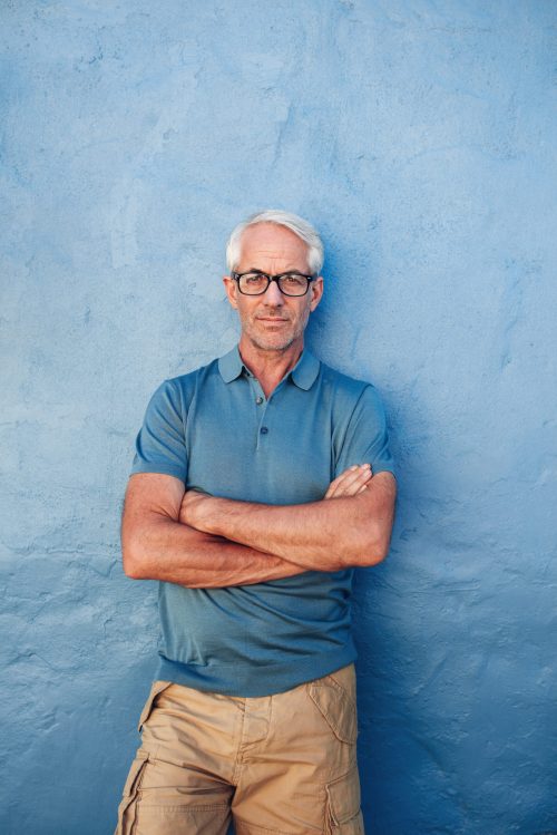 Vertical portrait of confident mature man standing against a blue background. Handsome caucasian man in glasses standing with his arms crossed against a wall.
